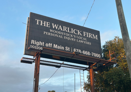 Outdoor billboard for The Warlick Firm advertising "Woodstock's Local Personal Injury Lawyers," featuring their location Right off Main St and phone number 678-668-7000.