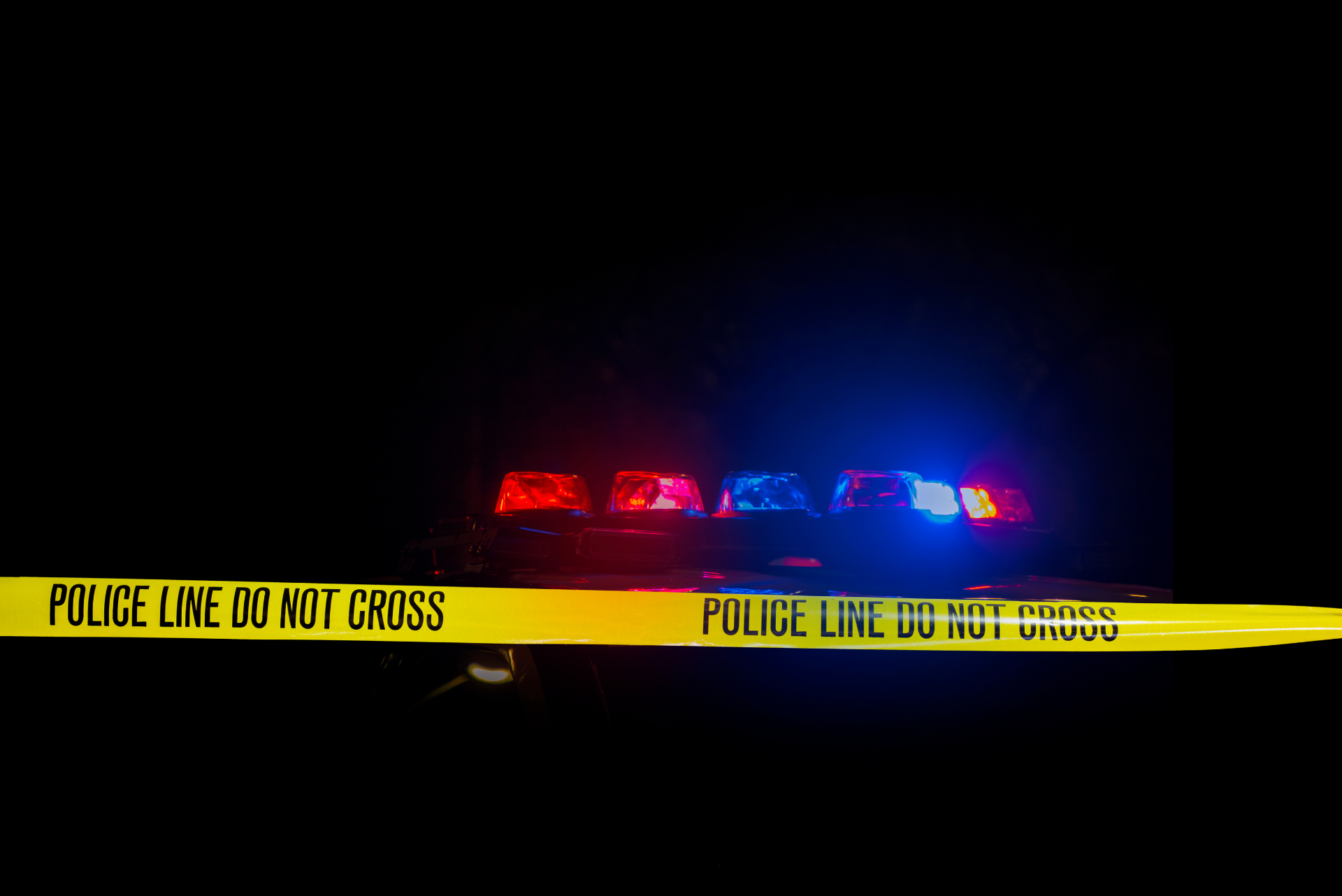 Yellow "Police Line Do Not Cross" tape stretched across the foreground with flashing red and blue police cruiser emergency lights glowing in the dark background.