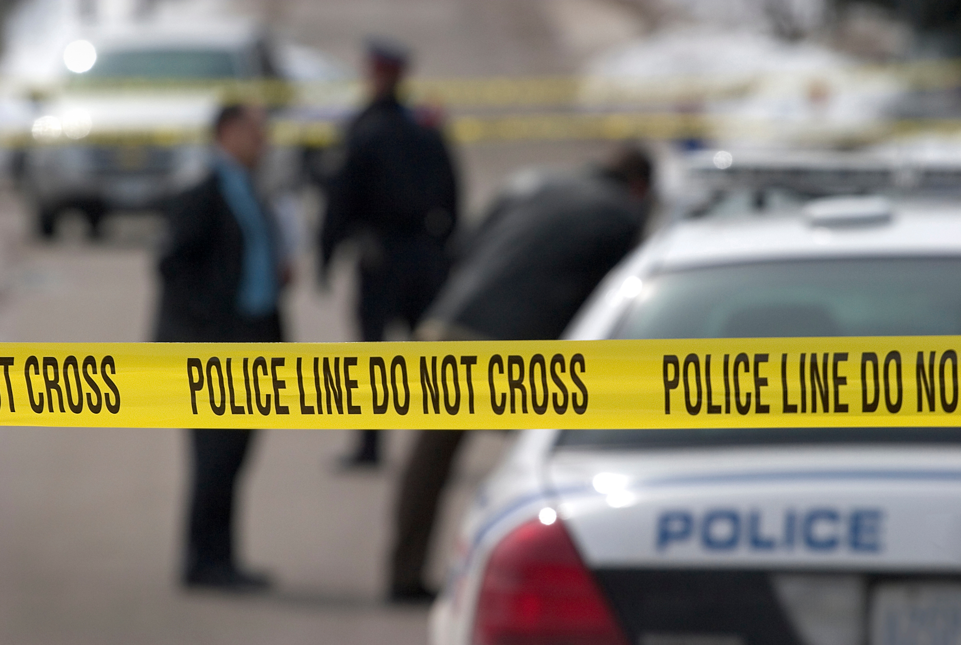 Yellow police tape reading "POLICE LINE DO NOT CROSS" stretched across an active crime scene with blurred police officers and a squad car in the background, illustrating a legal analysis regarding Galal Mohamed Abadi.